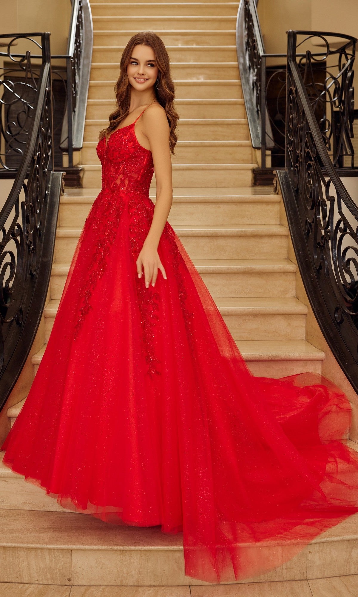 Wearing the Nox Anabel H1883, a long red prom dress, a woman stands on a staircase, smiling and posing with one hand slightly raised.